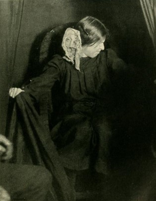 A picture containing a person dressed in black facing slightly away from the camera with something white next to her ear. 