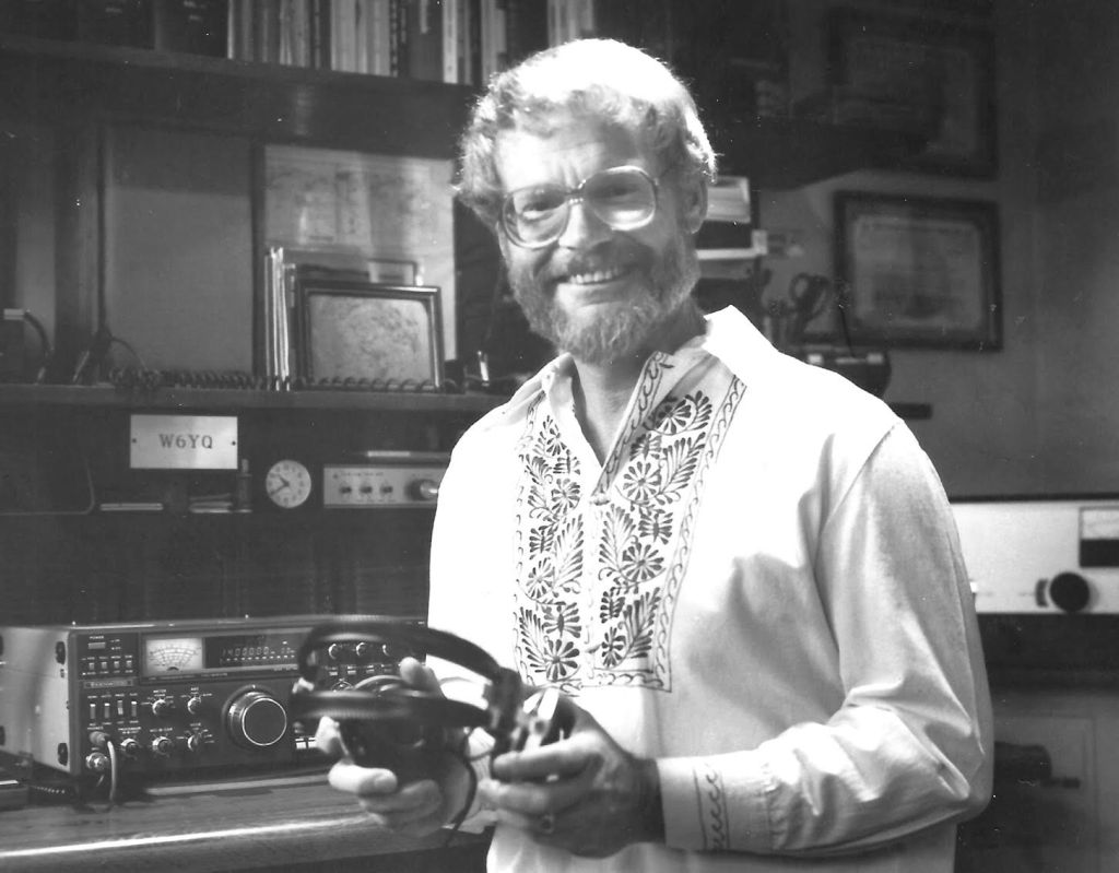 Black and white photo of Michael Hudgens holding headphones and standing next to a ham radio. 