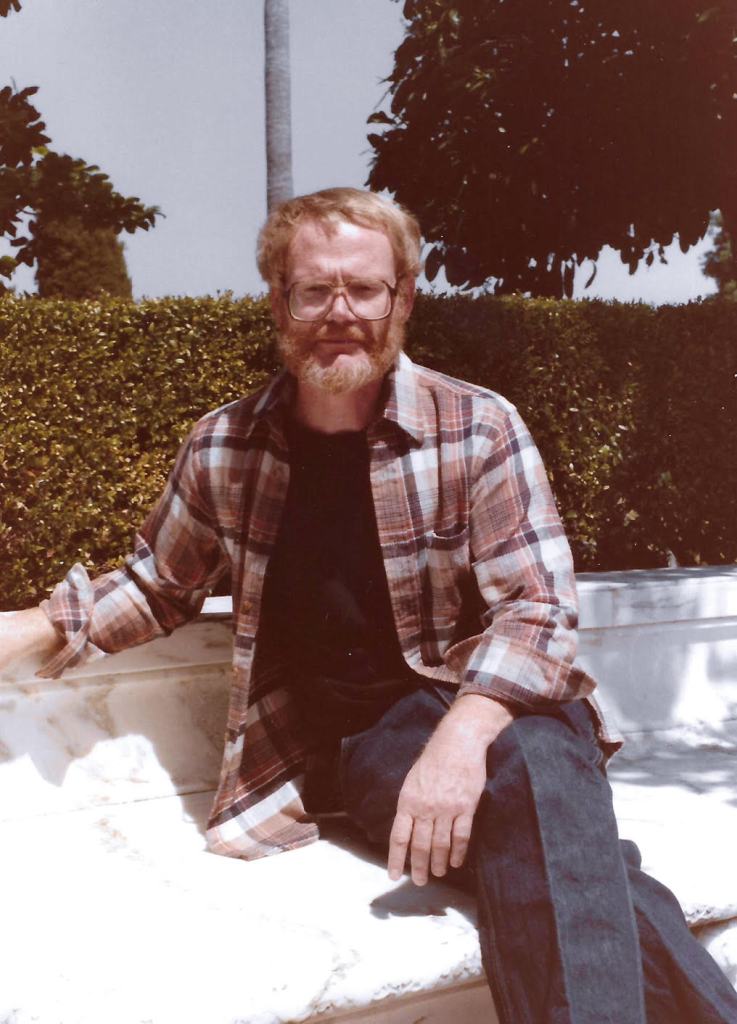 Michael Hudgens seated outside on a marble bench in front of bushes in the sunshine. 