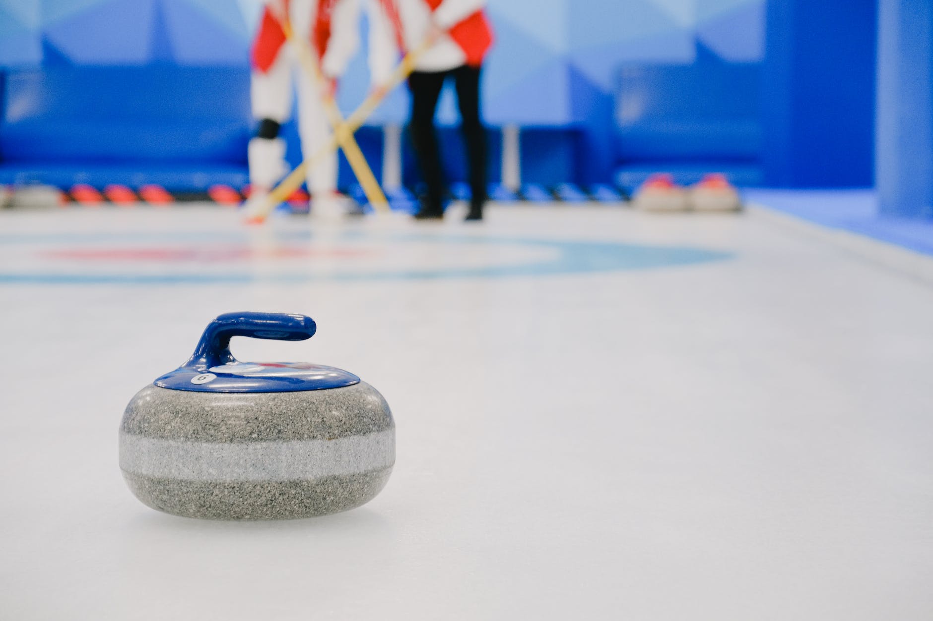 A curling stone sits on the ice in the foreground with people in the background holding brooms. 