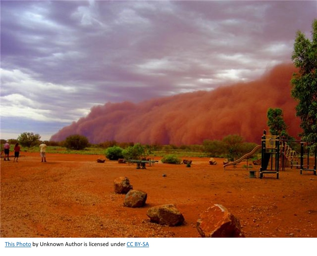 Photo of giant reddish-brown dust cloud blowing in from the right side of the image, approaching a playground and a few people watching it. 