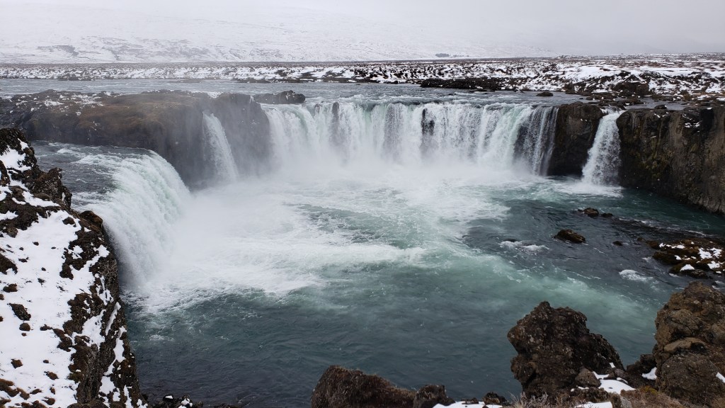 Photo of Godafoss waterfall in Iceland. Horseshoe-shaped falls in the center of the image, with snow-covered ground and rocks around it. 
