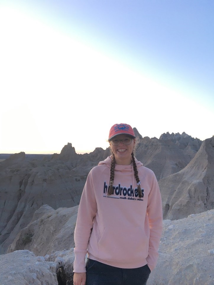 A young white woman in a pink sweatshirt and pink hat, with the Badlands behind her.