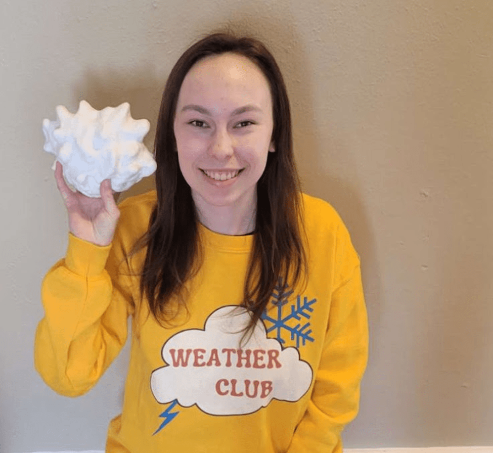 Young white woman with dark hair holding a 3-D printed hailstone model in one hand and wearing a bright yellow sweatshirt that says "Weather Club." 