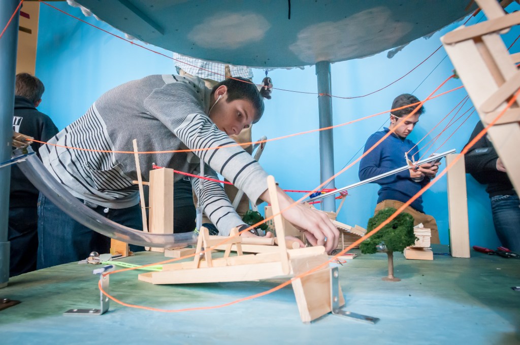 A young man adjusts a part on a Rube Goldberg machine. String and pieces of wood are in the foreground, as part of the machine. 