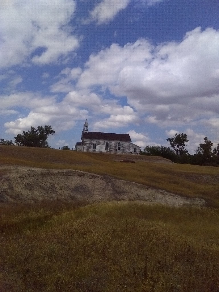 An old, abandoned church with peeling white paint on top of a hill with the blue sky and white clouds behind it. 