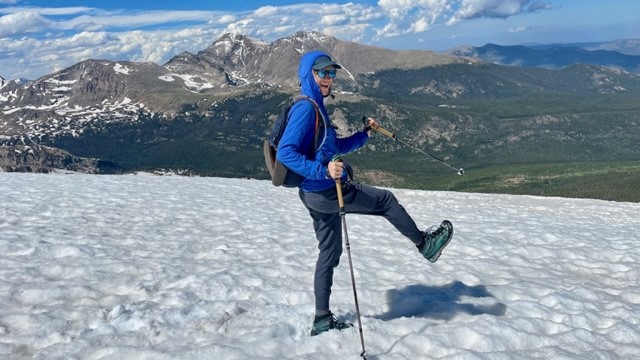 Mary hiking across a snow field with mountains and blue sky in the background. 