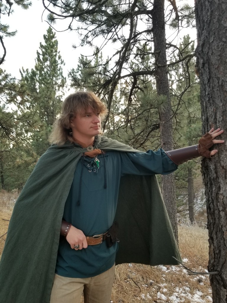 Young man posing in the forest with Renaissance-style clothing - a lace-up shirt, a cape, and leather wristbands. 