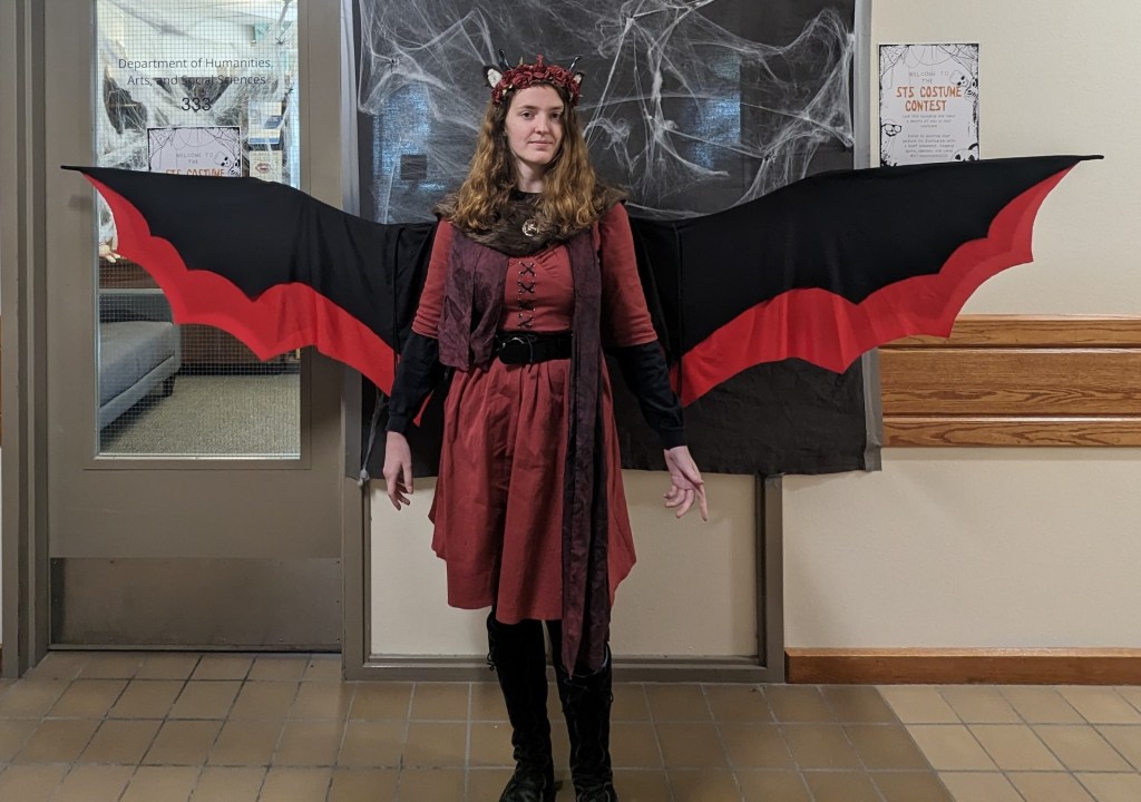 A young woman in a Renaissance-style lace-front dress with tall boots and a headpiece. Most notably, she is also wearing wide black-and-red wings. 