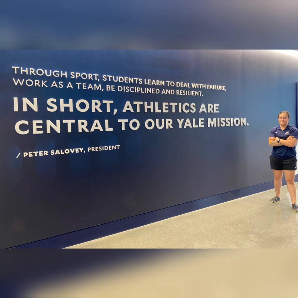 Sophia stands next to writing on a wall: "Through sport, students learn to deal with failure, work as a team, be disciplined and resilient. In short, athletics are central to our Yale mission."