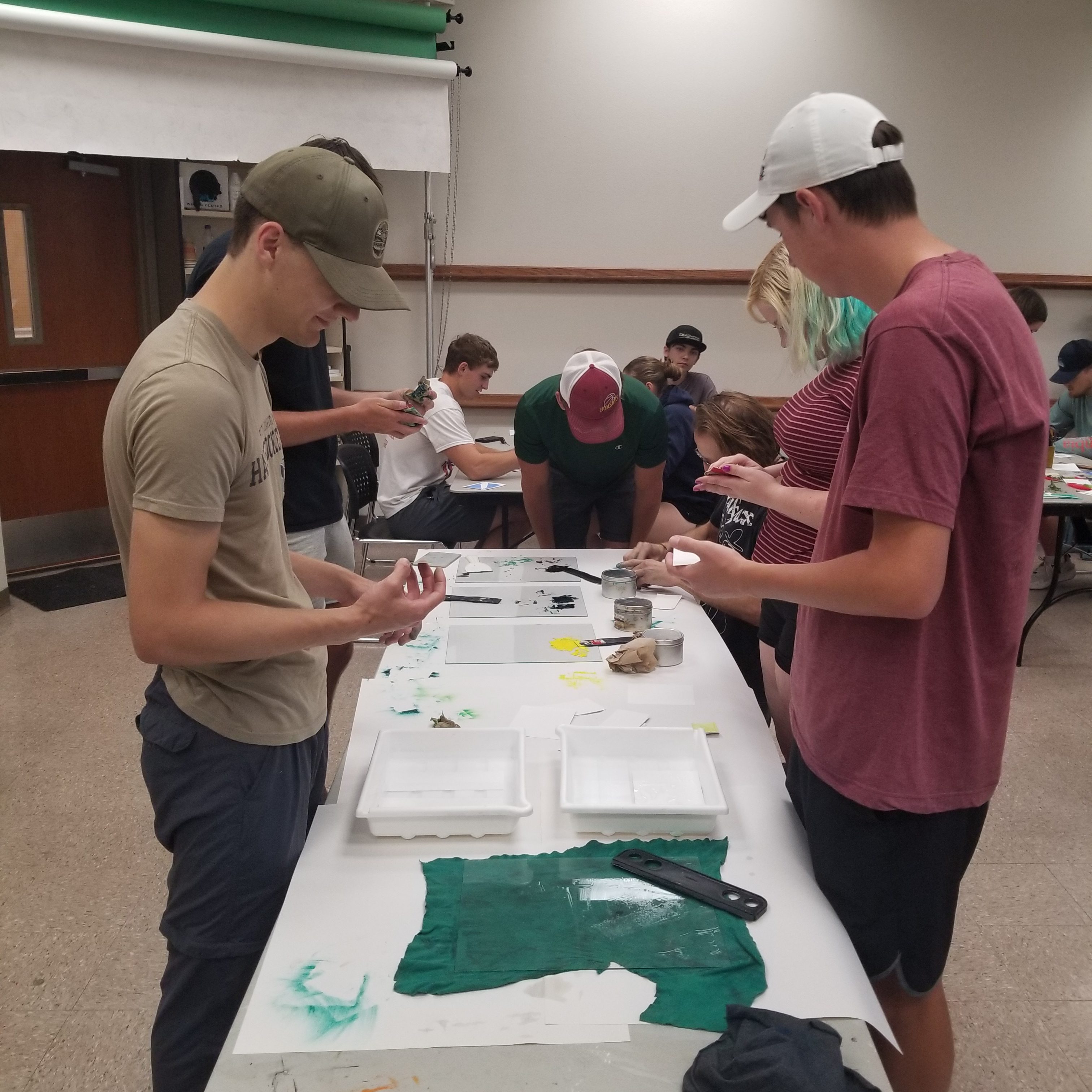 Students stand at a table applying ink in the foreground. In the background, more students sit and work on creating their own etchings. 
