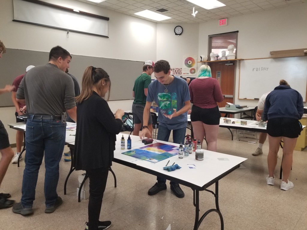 Students surround two tables working on their projects - applying ink. 