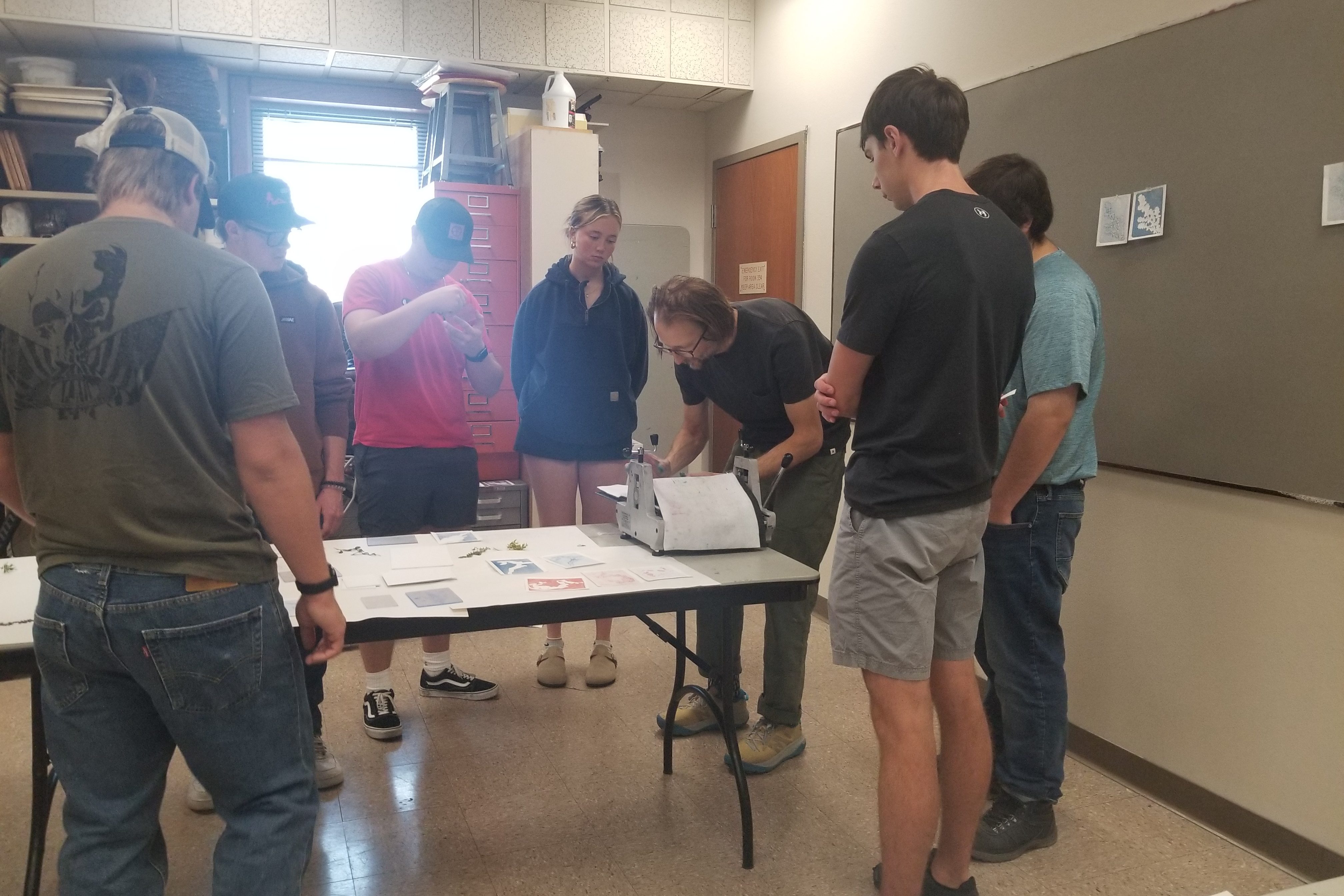 A man bends over a printing press while using it and a group of students stands around him, observing. 