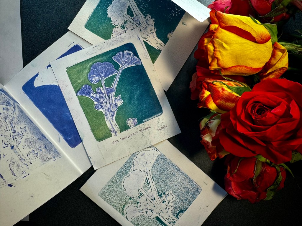 Photo of a set of prints, with the top  one including a green and blue printed plant and the text "with love, heart blooms." The prints are laid out next to some red and yellow roses. 