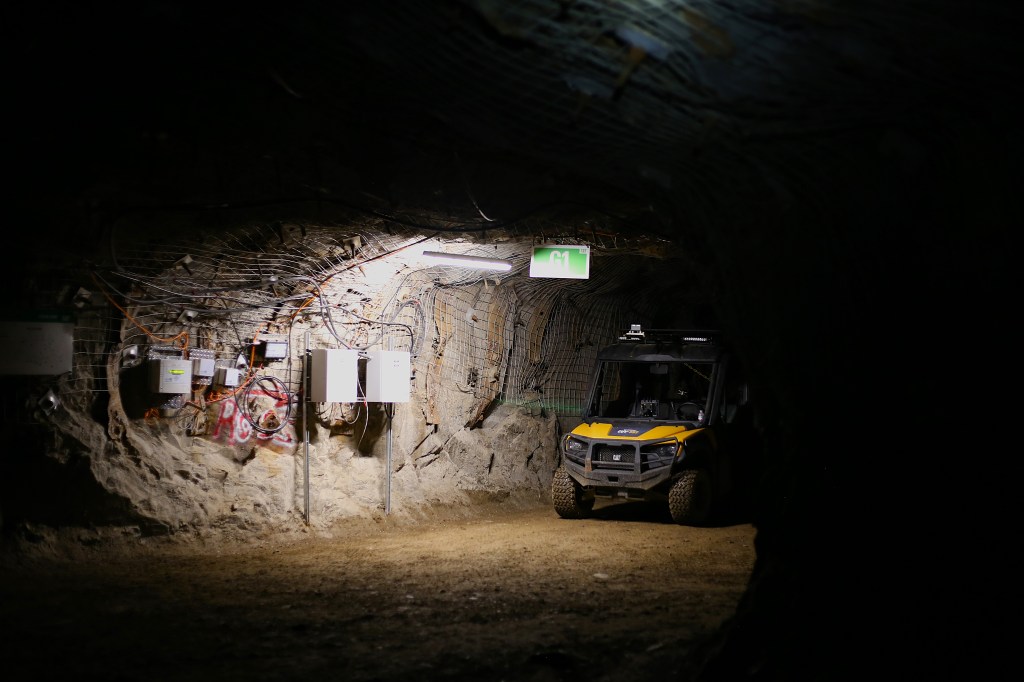A small vehicle sits in an underground space. A single bright light hangs from the ceiling and illuminates both the vehicle and the electrical wiring on the wall. 