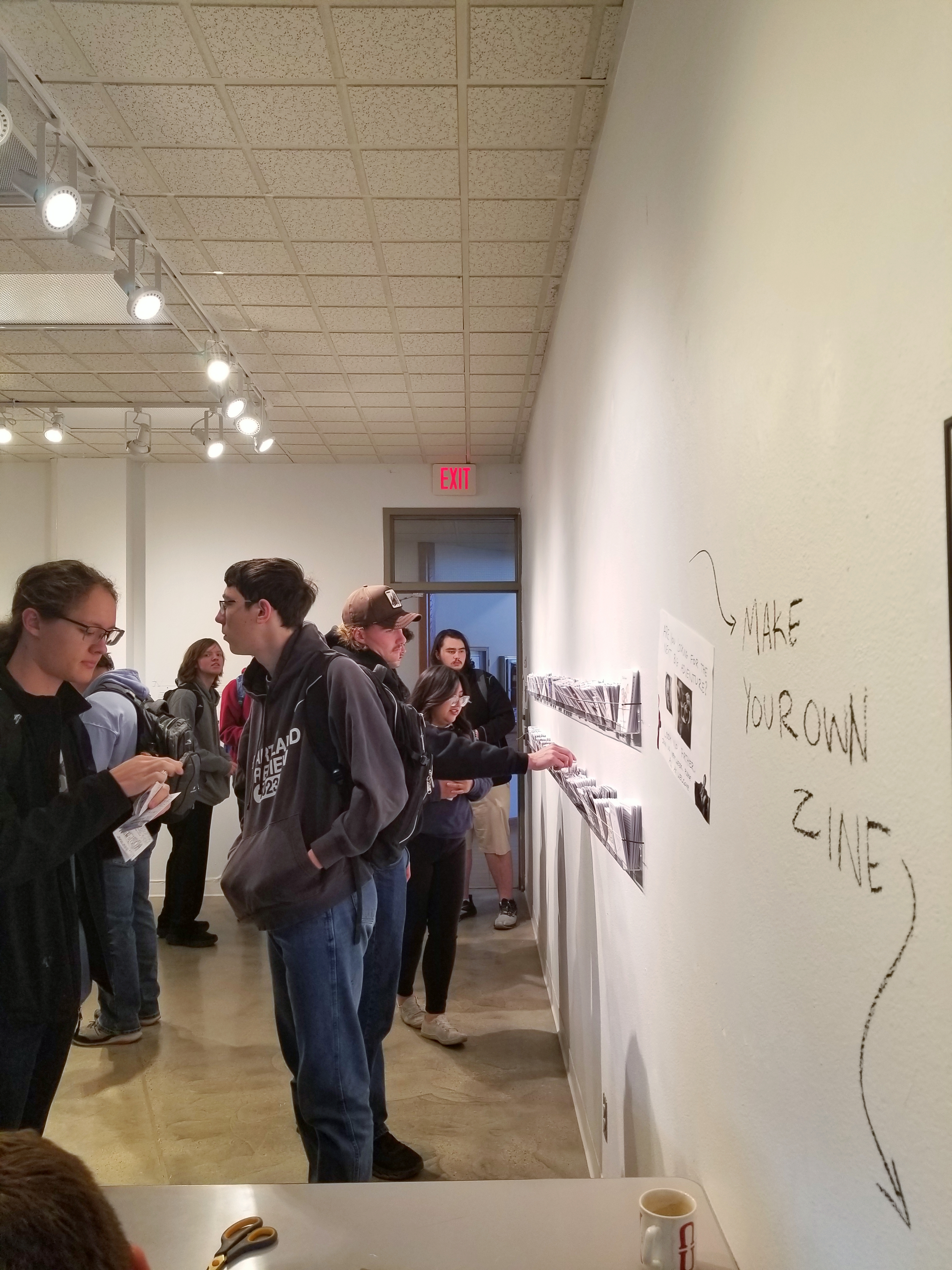 Zinefest attendees looking at zines. Writing on the wall points to a station to make your own zines. 