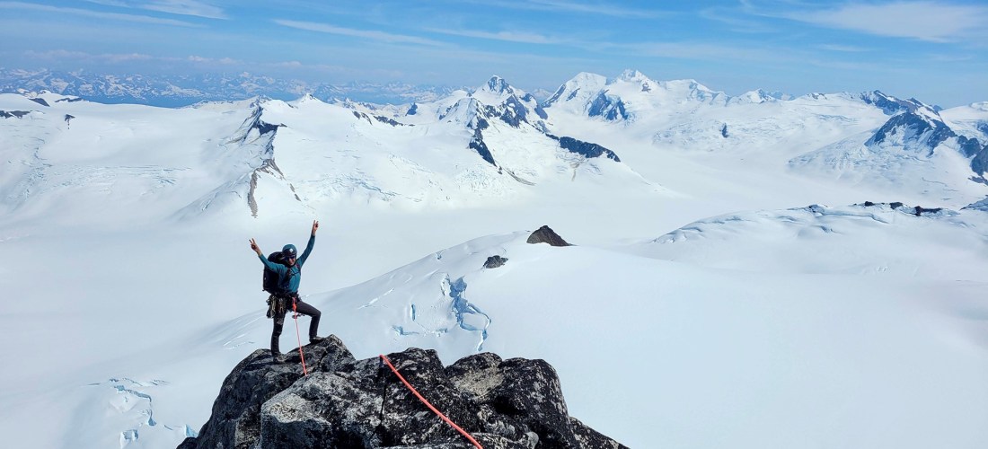 Climber on top of a peak, with snowy mountains extending into the background.