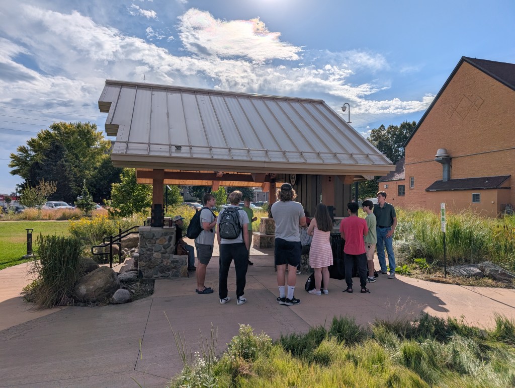 A group of students stands near a shelter at the park while they listen to the park director speak about it. 