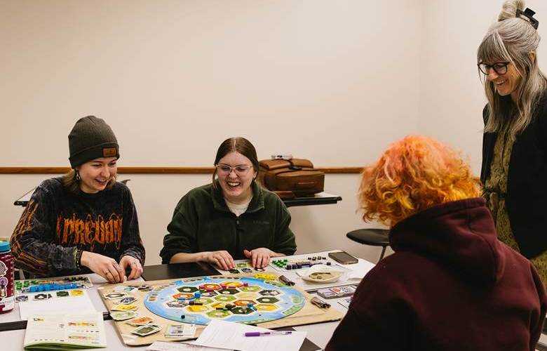 Students sit at a table playing the board game while Dr. Witlacil observes.