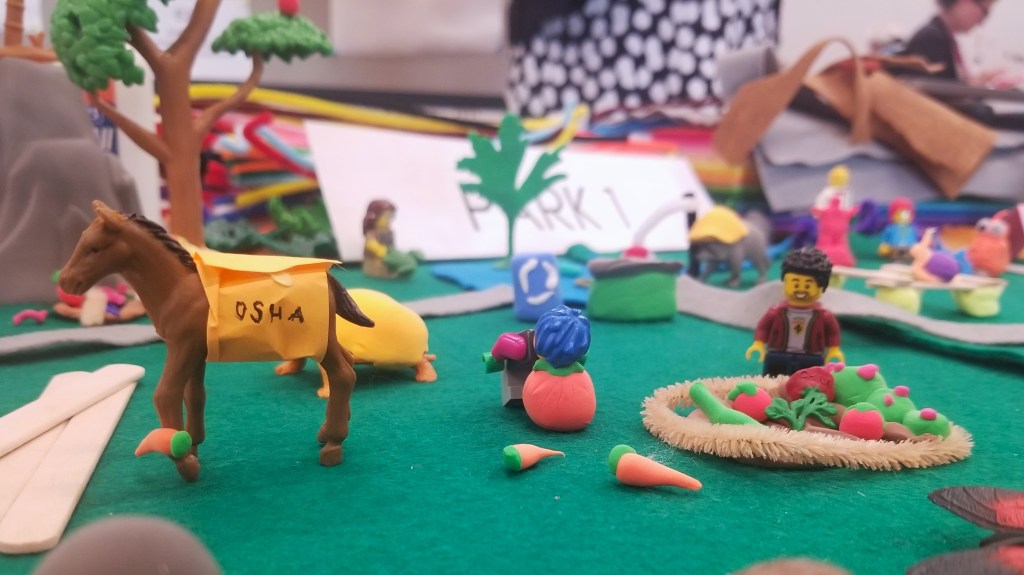 A detail shot from the Build-A-Park table that features a toy horse wearing a student-made OSHA vest and a small community garden with a Lego person standing next to it. 